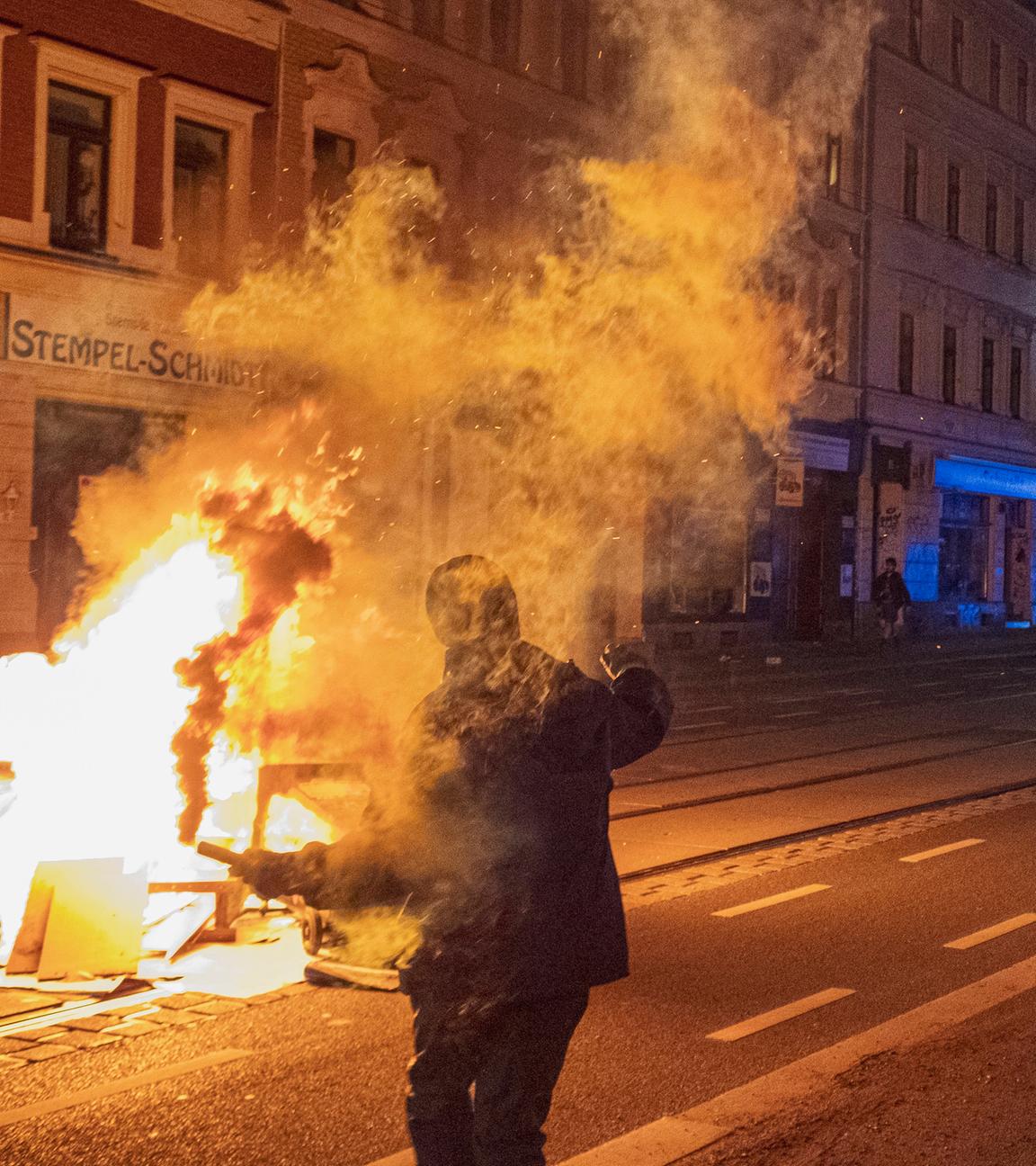 Sachsen, Leipzig: Ein maskerter Demonstrant steht mit brennender Pyrotechnik in der Hand auf einer Straße.