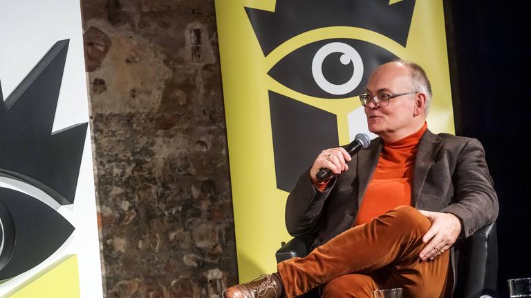 Ein Mann mittleren Alters sitzt auf einem Podium mit dem Logo der Leipziger Buchmesse im Hitergrund.