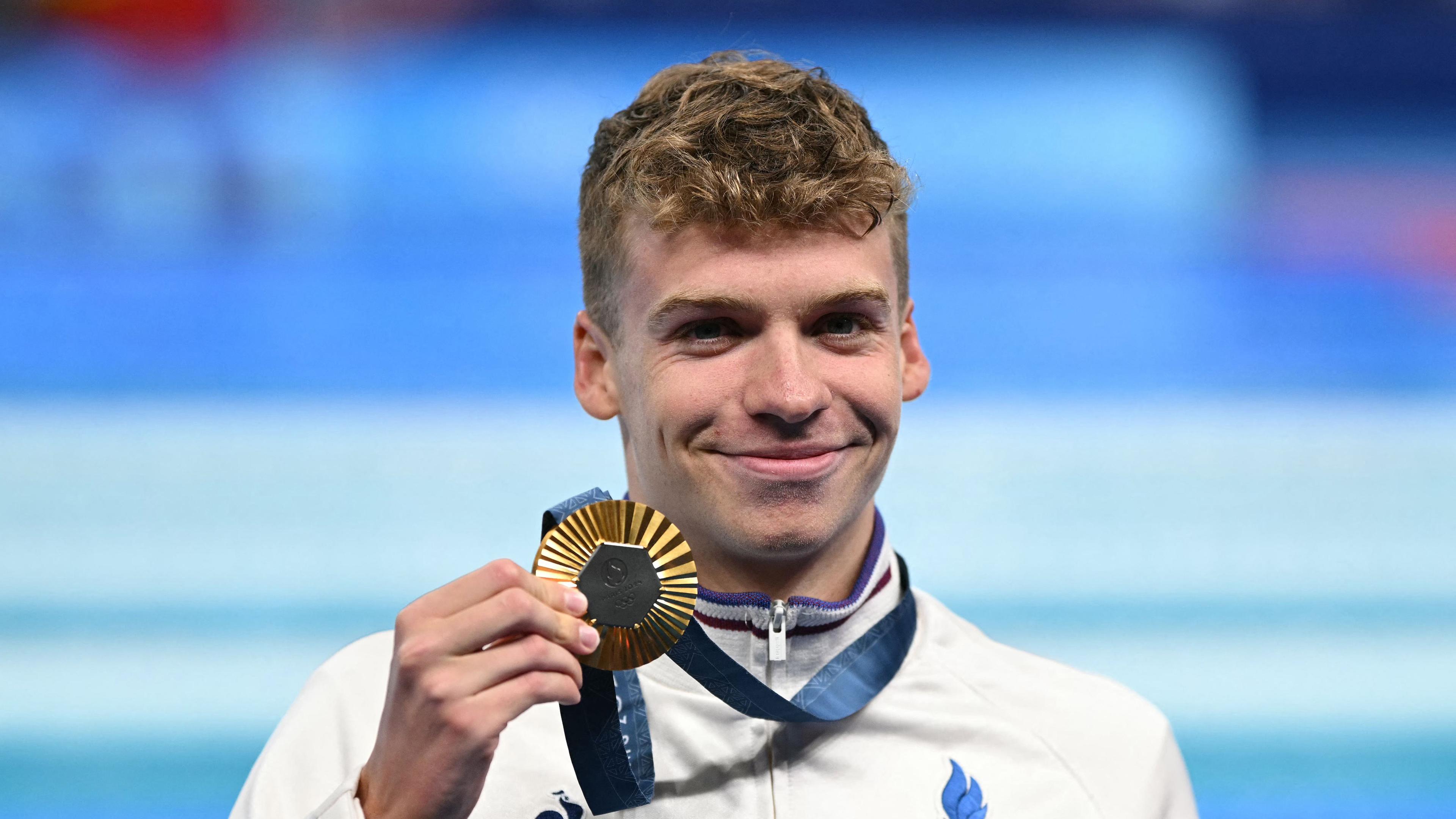 León Marchand hält eine Goldmedaille in der Hand und lacht.