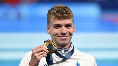 León Marchand hält eine Goldmedaille in der Hand und lacht.