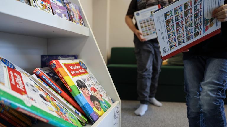 Links ist ein Regal mit Lernbüchern. Rechts stehen zwei Kinder mit Büchern in der Hand.