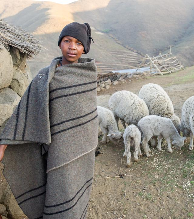 Zwei junge Schafhirten stehen in einer Decke gewickelt neben ihrer Herde in einer Bergregion im afrikanischen Lesotho.