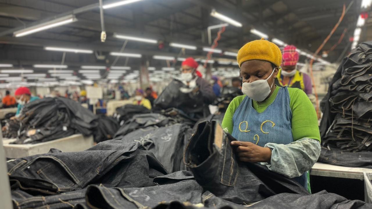 Eine Frau steht in einer Fabrikhalle und ist mit einem großen Stapel Jeans beschäftigt.