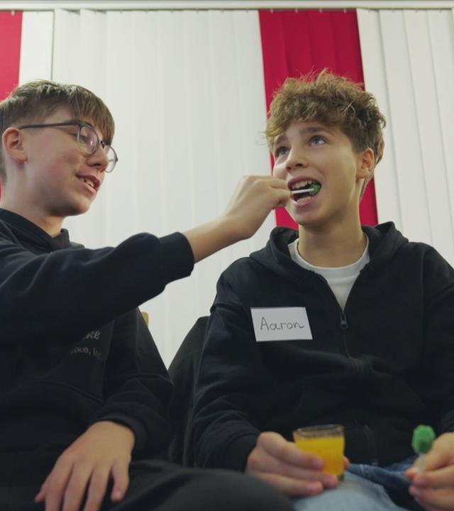 Drei Kinder sitzen nebeneinander. Zwei Jungs üben, wie man sich gegenseitig den Mundraum reinigt.