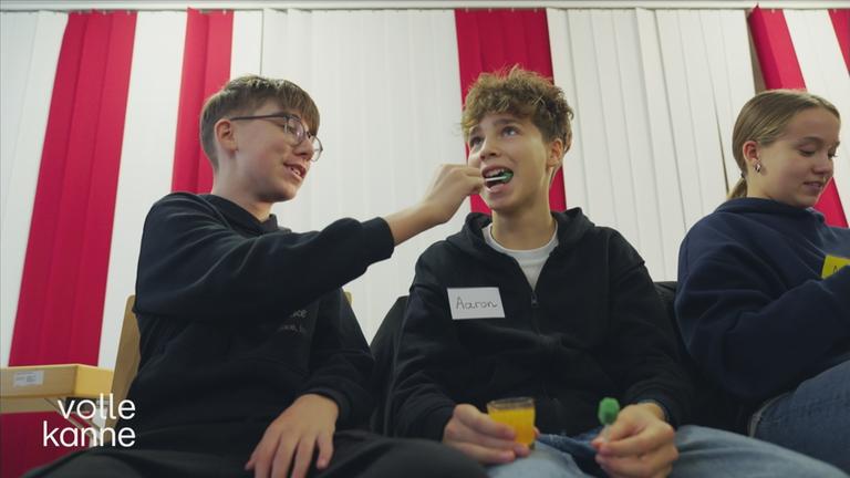 Drei Kinder sitzen nebeneinander. Zwei Jungs üben, wie man sich gegenseitig den Mundraum reinigt.