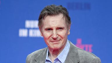 Liam Neeson posiert vor einem blauen Hintergrund auf einem Event.