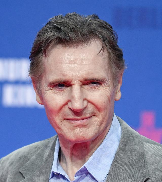 Liam Neeson posiert vor einem blauen Hintergrund auf einem Event.