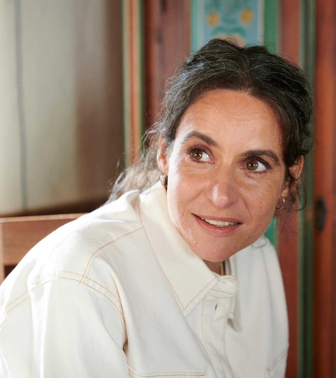 Schauspielerin Liane Forestieri sitzt in häuslicher Umgebung vor einem altem Schrank mit schweifendem Blick nach links.