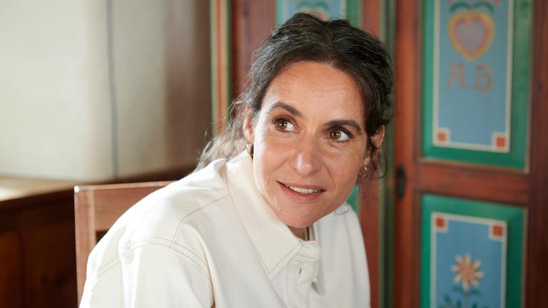 Schauspielerin Liane Forestieri sitzt in häuslicher Umgebung vor einem altem Schrank mit schweifendem Blick nach links.