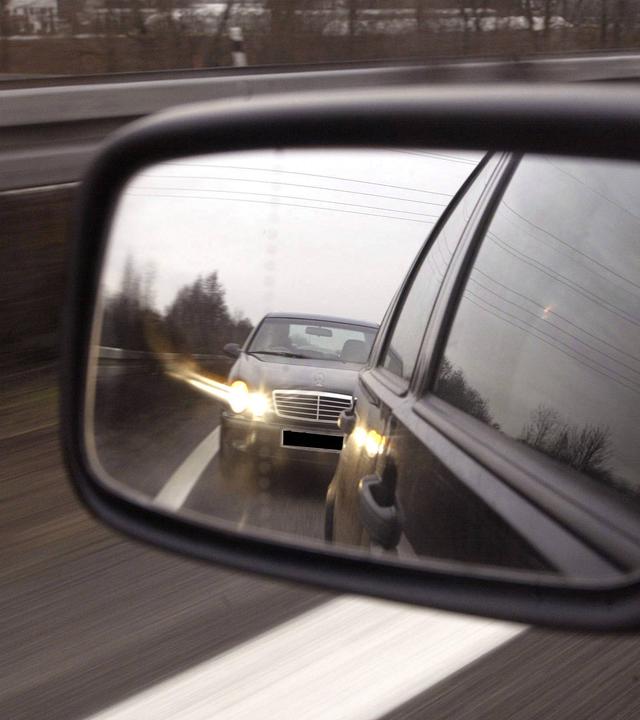 Im Außenspiegel eines Pkw ist ein Drängler zu sehen, der ganz dicht aufgefahren ist und per Lichthupe die Freigabe der Fahrspur fordert.