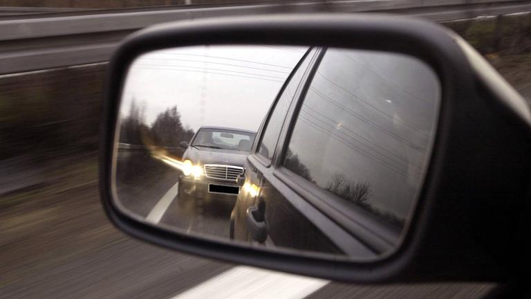 Im Außenspiegel eines Pkw ist ein Drängler zu sehen, der ganz dicht aufgefahren ist und per Lichthupe die Freigabe der Fahrspur fordert.