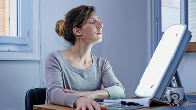 Eine Frau schaut mit geschlossenen Augen in Richtung einer Tageslichtlampe auf einem Tisch.