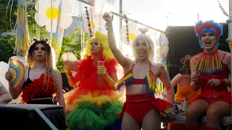 Vier Dragqueens feiern auf einem Festwagen die Pride Parade. Sie tragen bunte Perücken und Kostüme.