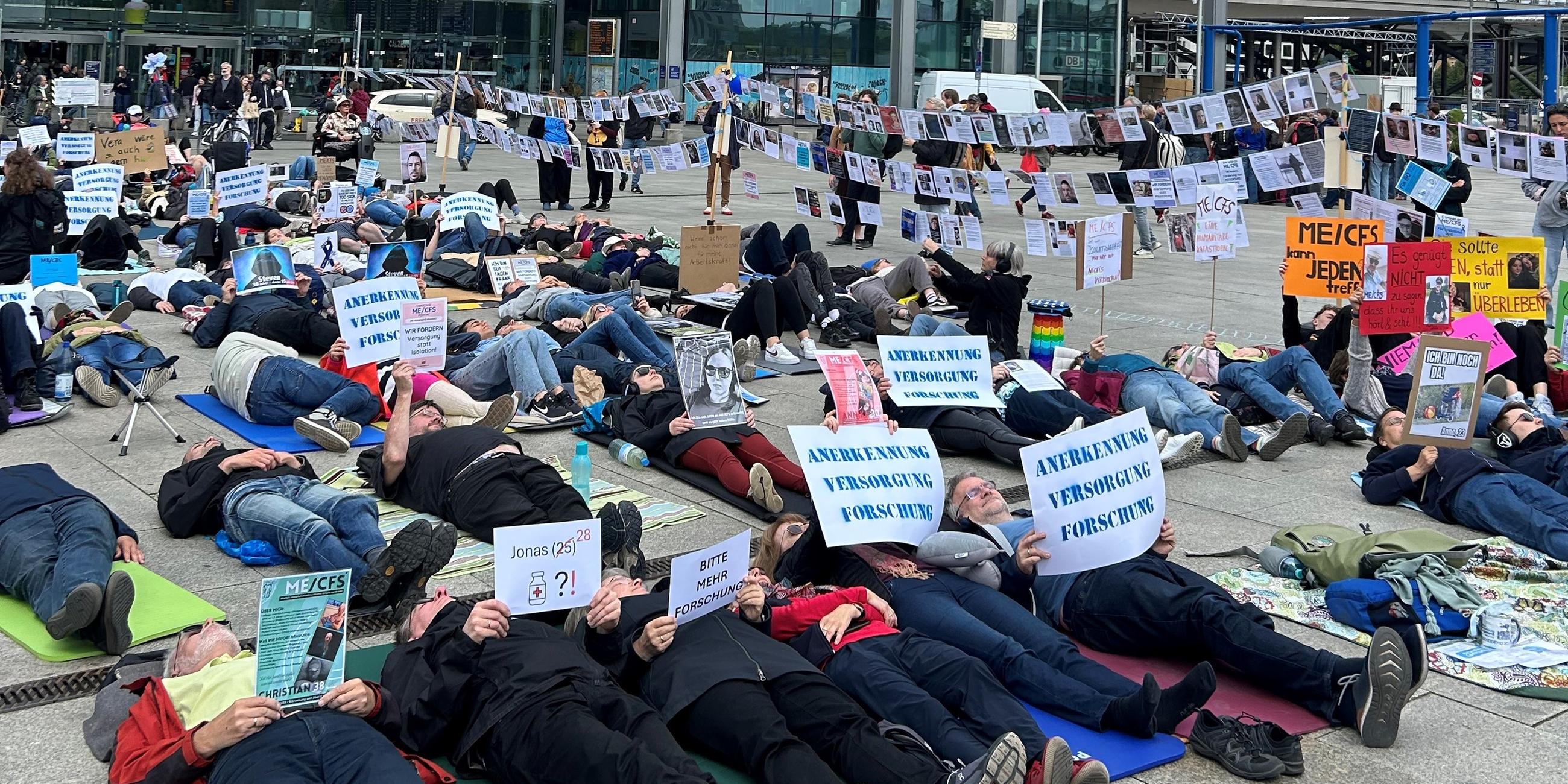Viele Menschen liegen auf dem Boden bei einer Liegenddemo in Berlin, die auf die Krankheit ME/CFS aufmerksam machen soll. Auf ihren Plakaten steht "Anerkennung - Versorgung - Forschung".