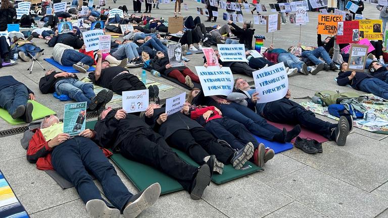 Viele Menschen liegen auf dem Boden bei einer Liegenddemo in Berlin, die auf die Krankheit ME/CFS aufmerksam machen soll. Auf ihren Plakaten steht "Anerkennung - Versorgung - Forschung".