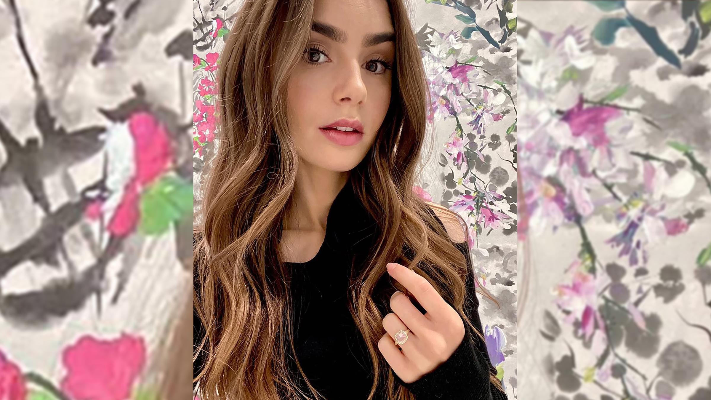 Lily Collins schaut in die Kamera und hält ihre Hand mit dem Verlobungsring am Ringfinger hoch.