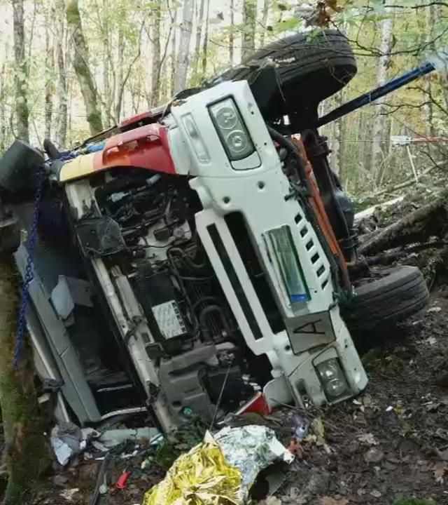 Auf dem Bild ist zu sehen: Ein stark beschädigter Lkw liegt seitlich an einer bewaldeten Böschung. Seine weiße Fahrzeugfront ist zur Kamera gerichtet. Links unten ist eine Einsatzkraft zu sehen.