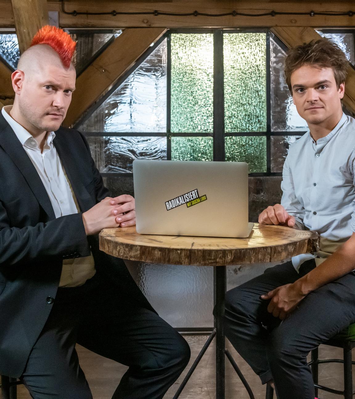 Sascha Lobo und Patrick Stegemann sitzen gemeinsam an einem Tisch, auf dem ein aufgeklappter Laptop steht.