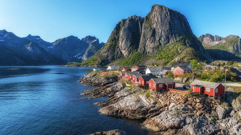 Ein kleiner Ort am Wasser. Die roten Häuser stehen fast direkt am Wasser. Im Hintergrund sind hohe Berge, die ein wenig bewachsen sind.