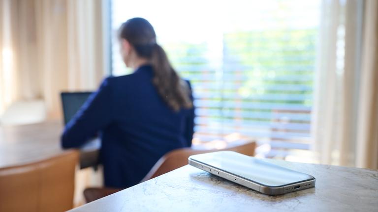 Ein Smartphone liegt auf einem Tisch, im Hintergrund arbeitet eine Frau an einem Laptop.
