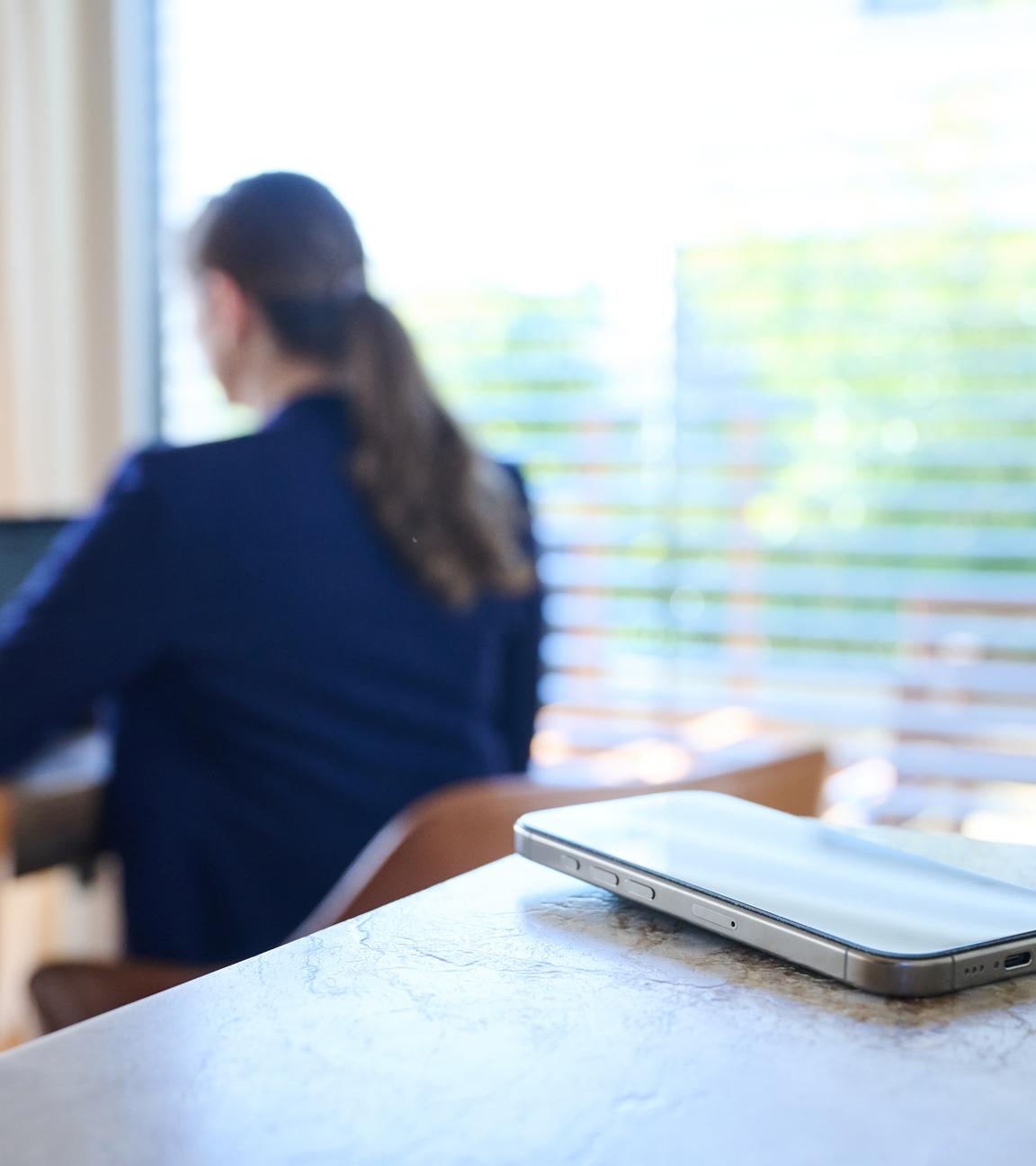 Symbolbild: Ein Smartphone liegt auf einem Tisch, im Hintergrund arbeitet eine Frau an einem Laptop.