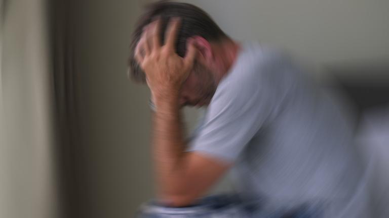 Auf dem Foto ist ein Mann zu sehen, der auf einer Bettkante sitzt. Beide Arme halten seinen Kopf. 