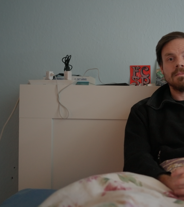 Fabian Fritz sitzt aufrecht im seinem Bett. Seine Hände sind über der Decke gefaltet, die über seinen Beinen liegt. Auf dem Kopfteil des Bettes stehen einige Grußkarten, eine Zeichnung von Asterix dem Gallier. Daneben liegen zwei Bücher und eine Steckdosenleiste, in der zwei Ladekabel eingesteckt sind. Fabian trägt eine dunkle Fliesjacke mit einem Reißverschluss vorne. Er trägt einen dunkle Bart, das Har ist kurz geschnitten. Fabian blickt in die Kamera, die fronalt auf ihn gerichtet ist.