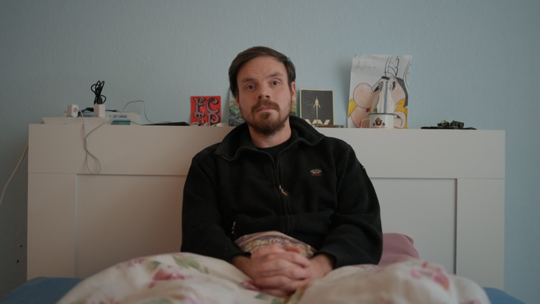 Fabian Fritz sitzt aufrecht im seinem Bett. Seine Hände sind über der Decke gefaltet, die über seinen Beinen liegt. Auf dem Kopfteil des Bettes stehen einige Grußkarten, eine Zeichnung von Asterix dem Gallier. Daneben liegen zwei Bücher und eine Steckdosenleiste, in der zwei Ladekabel eingesteckt sind. Fabian trägt eine dunkle Fliesjacke mit einem Reißverschluss vorne. Er trägt einen dunkle Bart, das Har ist kurz geschnitten. Fabian blickt in die Kamera, die fronalt auf ihn gerichtet ist.