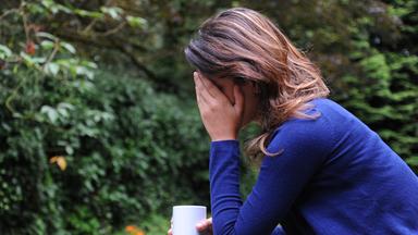 Eine Frau sitzt mit einer Kaffeetasse in der Hand in einem Garten. Sie stützt sich erschöpft auf einer Hand ab.