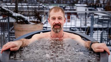 Daniel sitzt auf seiner Dachterrasse beim Eisbaden in einer Tonne mit Wasser. Im Hintergrund liegt eine winterliche Stadtlandschaft.