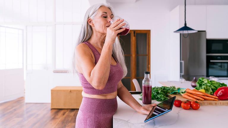 Eine Frau mit grauen Haaren hält in der einen Hand ein Tablet und trinkt aus einem Glas Saft in einer Küche.
