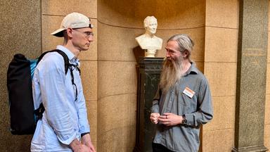 In der Innenwand eines Gebäudes steht eine Büste in einer Nische. Davor stehen Felix und Aubrey de Greay und unterhalten sich.