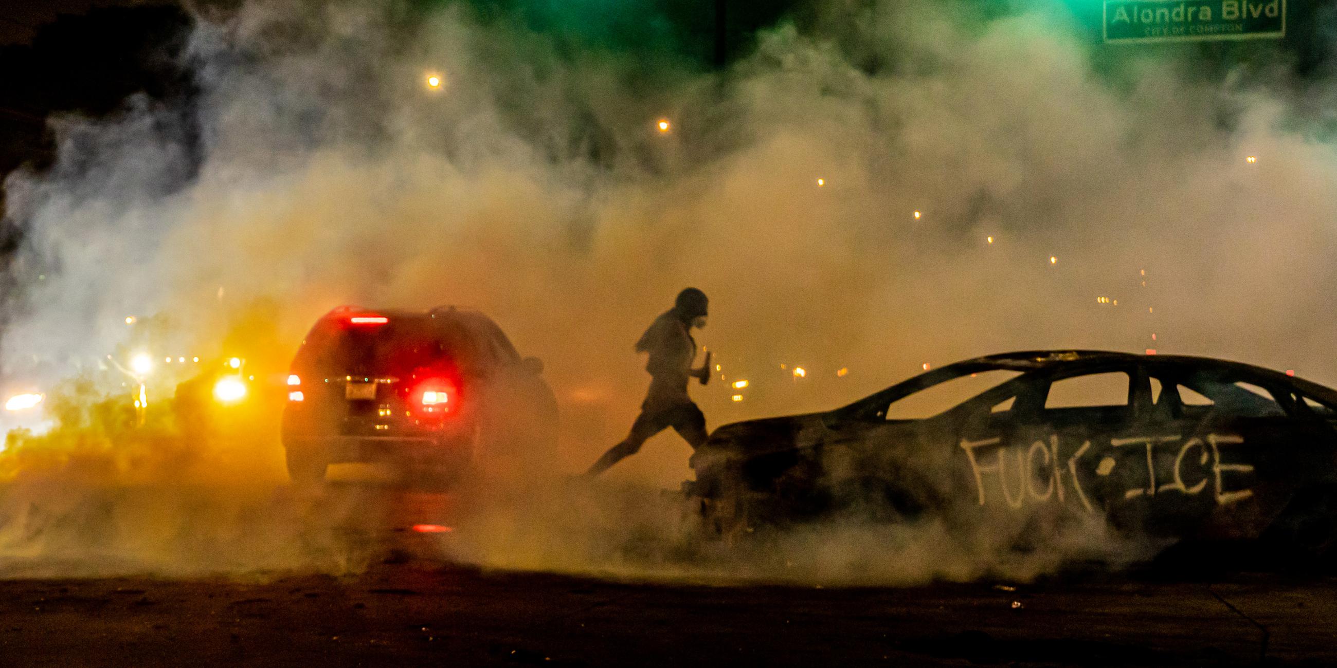 Zwei Autos bei Nacht an einer grünen Ampel, auf dem einen ist "Fuck ICE" gesprayt, um die Autos herum Rauch, in der Mitte läuft eine Person durch.