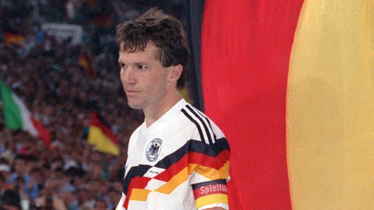 Lothar Matthaeus im Deutschandtrikot vor einer Deutschland-Flagge und erwartet die Siegerehrung 1990.