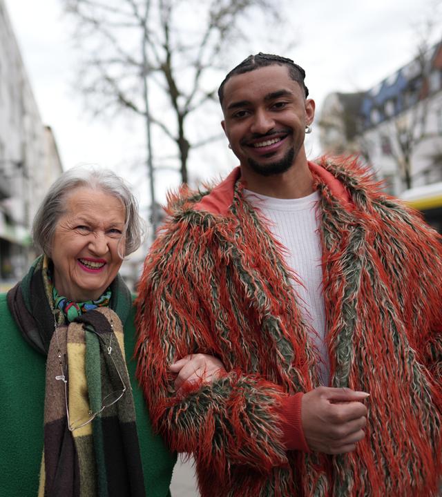 Louis (rechts) und seine Oma (links) schauen lächelnd in die Kamera. Sie hat sich bei ihm am Arm eingehakt. Im Hintergrund sieht man einen Baum und eine Straße.