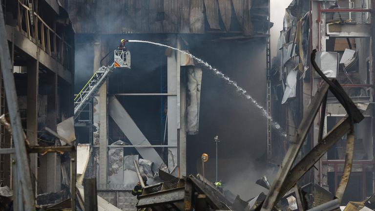 Ein zerstörtes Gebäude und eine Feuerwehrleiter, von der aus eine Person mit Wasser einen Brand zu löschen versucht.