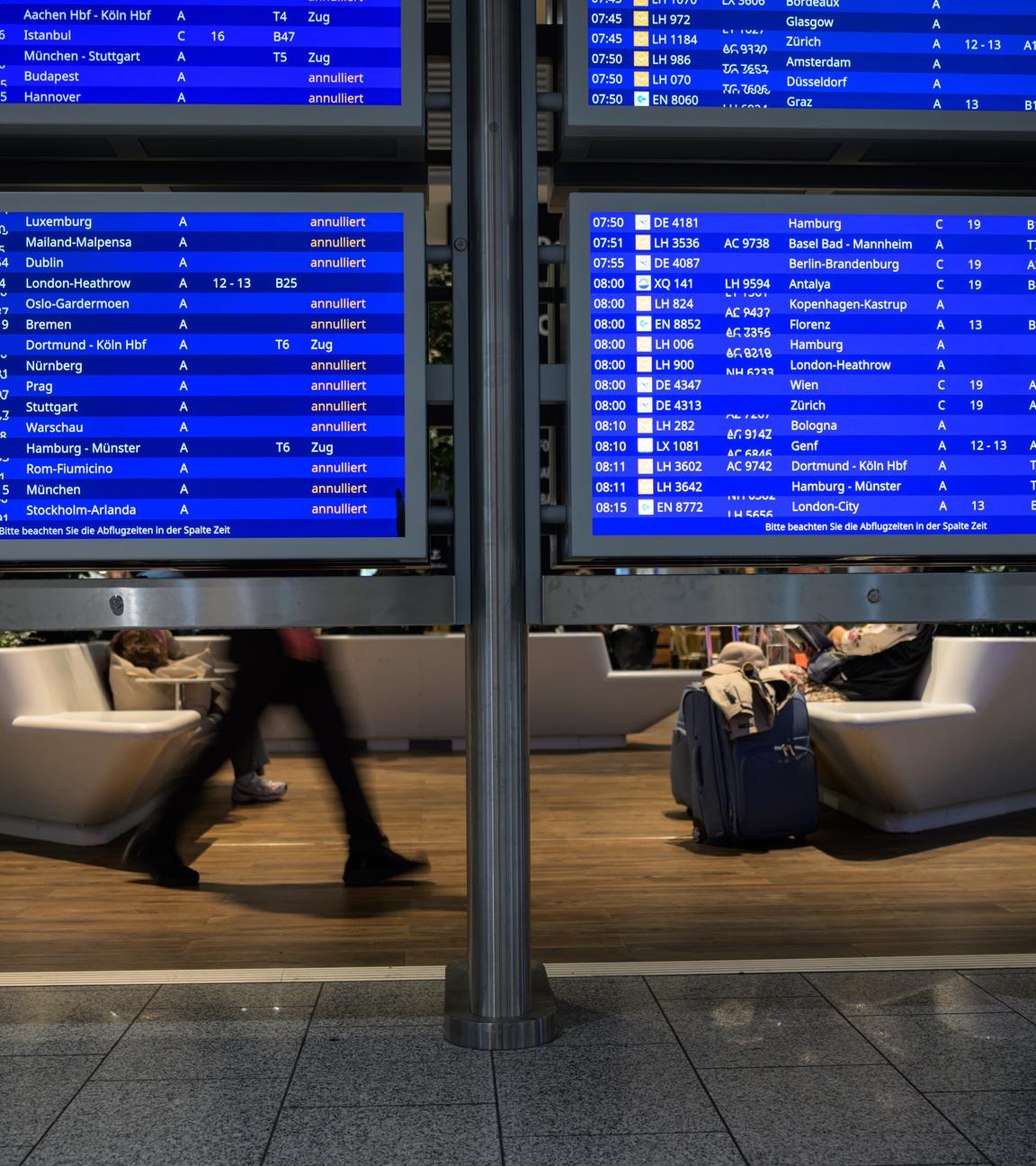16.04.2026, Hessen, Frankfurt/Main: Menschen sitzen in einem Wartebereich hinter Anzeigetafeln mit annullierten Flügen am Frankfurter Flughafen.