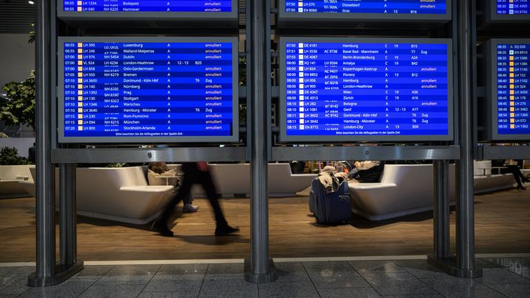 16.04.2026, Hessen, Frankfurt/Main: Menschen sitzen in einem Wartebereich hinter Anzeigetafeln mit annullierten Flügen am Frankfurter Flughafen.