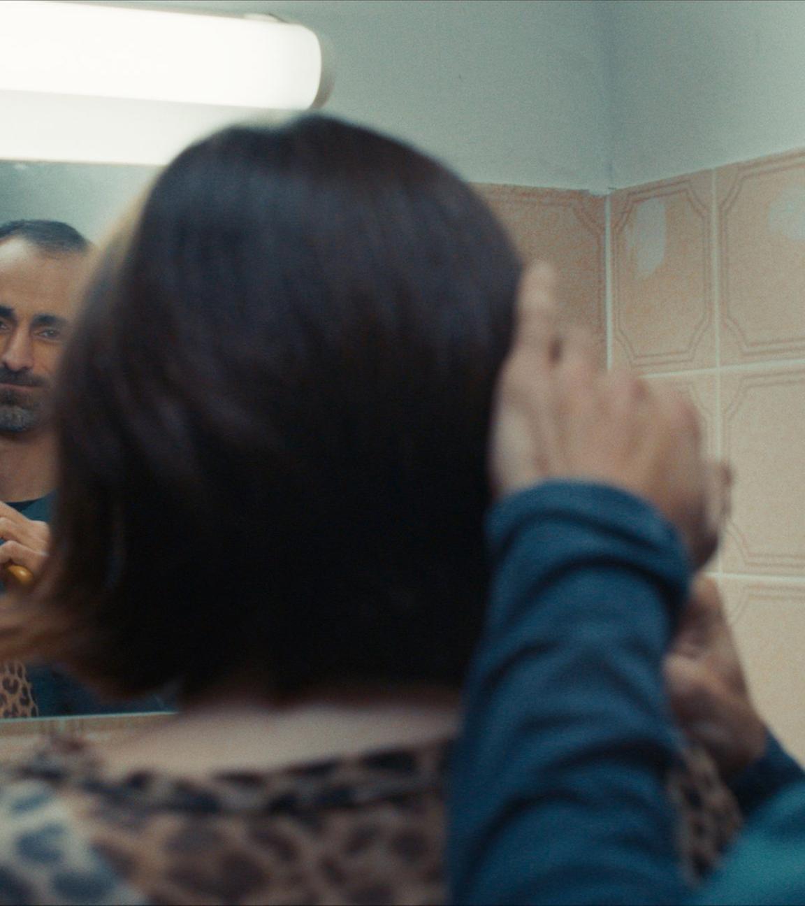 Ein Mann kämmt einer jungen Frau die Haare vor einem Badspiegel. Man sieht die Personen von hinten und  ihr Spiegelbild´.