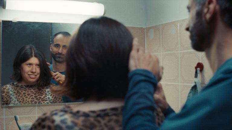 Ein Mann kämmt einer jungen Frau die Haare vor einem Badspiegel. Man sieht die Personen von hinten und  ihr Spiegelbild´.