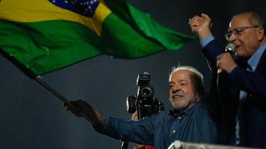 Lula da Silva hält eine Brasilien-Fahne in die Luft und jubelt. Neben ihm spricht ein anderer Mann ins Mikrofon.