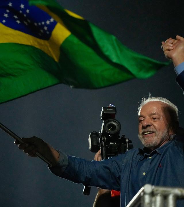 Lula da Silva hält eine Brasilien-Fahne in die Luft und jubelt. Neben ihm spricht ein anderer Mann ins Mikrofon.
