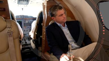Julien Backhaus sitzt mit überschlagenen Beinen in einem Privatjet mit einer Wasserflasche in der Hand. Er schaut aus dem Fenster. Im Hintergrund sind die Schultern von Pilot und Co-Pilot im Cockpit zu sehen.