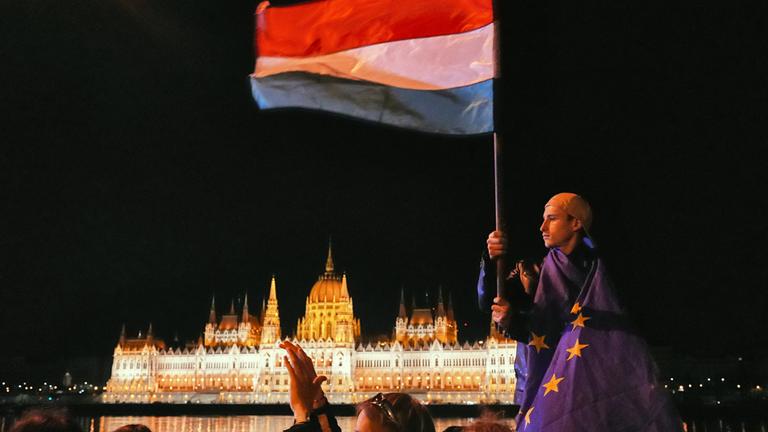Ungarn: Budapest: Ein Mann ist in die Flagge der EU gehüllt und schwenkt die ungarische Flagge vor dem Parlamentsgebäude.