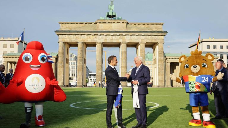 French President Emmanuel Macron and German President Frank-Walter Steinmeier shake hands as they exchance football shirts next to "Albaert" the official mascot of the UEFA Euro 2024 European Football Championship (R) and "Phryge" the Paris 2024 Olympic mascot, as they inaugurate the German-French Sports Summer 2024 in front of the Brandenburg Gate in Berlin, Germany on May 26, 2024. 