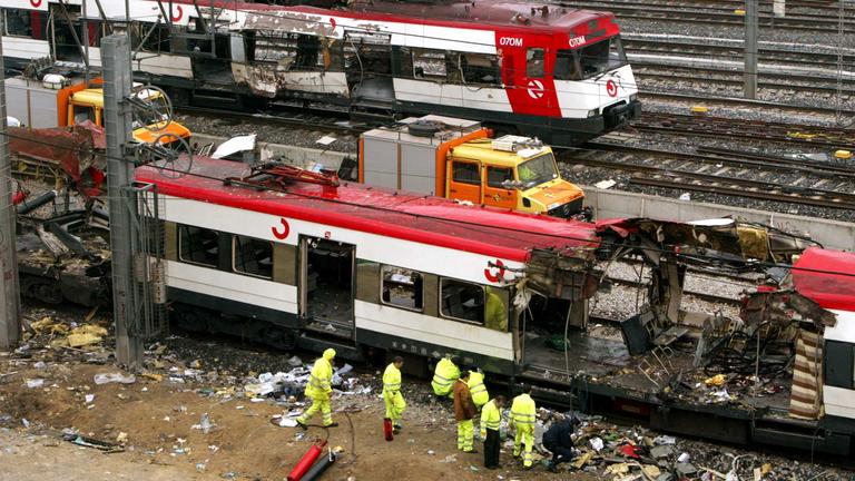 Menschen in gelben Warnjacken stehen neben einem Zugwagon, der durch eine Bombe zerstört wurde
