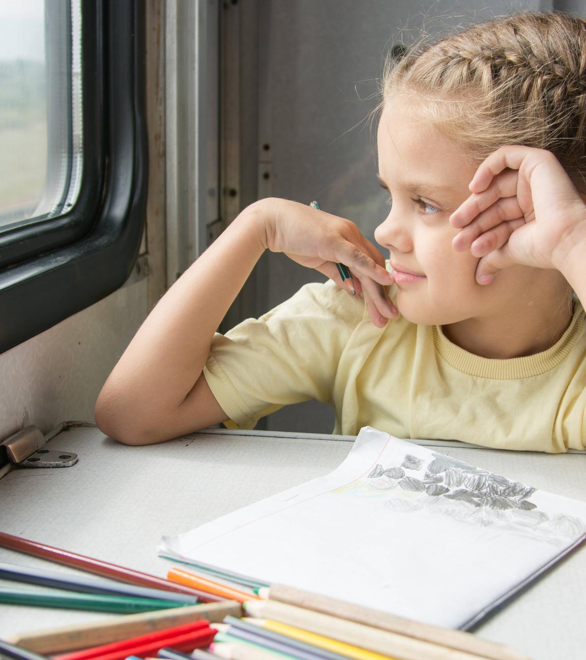Ein Mädchen sitzt während einer Bahnreise in einem Zugabteil am Tisch und malt.