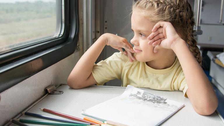 Ein Mädchen sitzt während einer Bahnreise in einem Zugabteil am Tisch und malt.