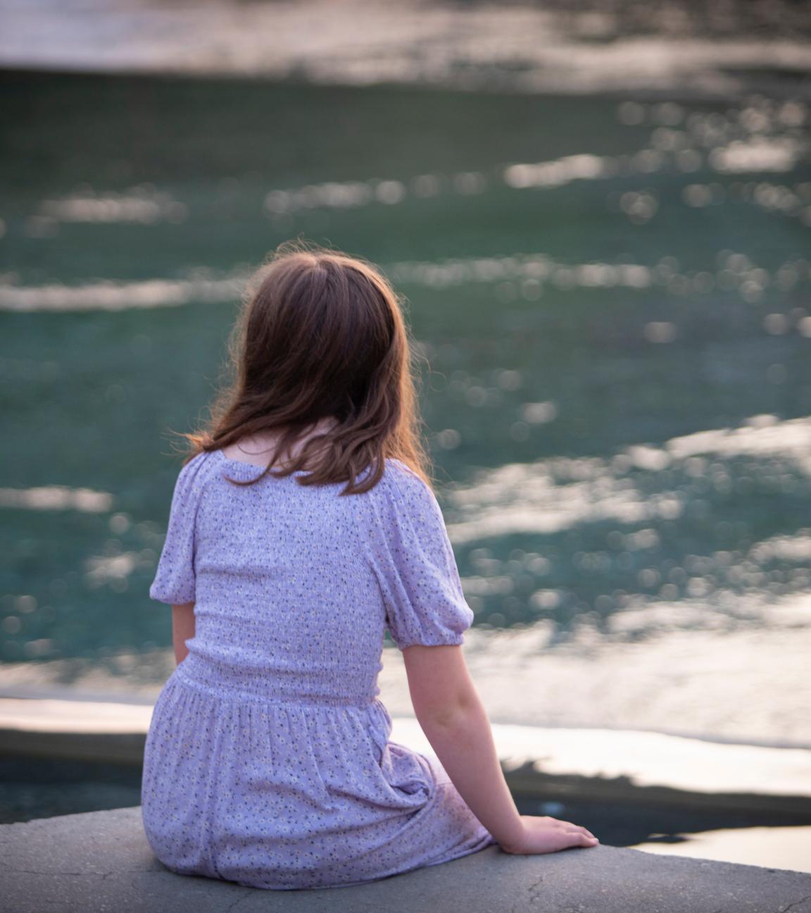 Ein Mädchen sitzt mit dem Rücken zur Kamera an einem Fluss-Ufer.