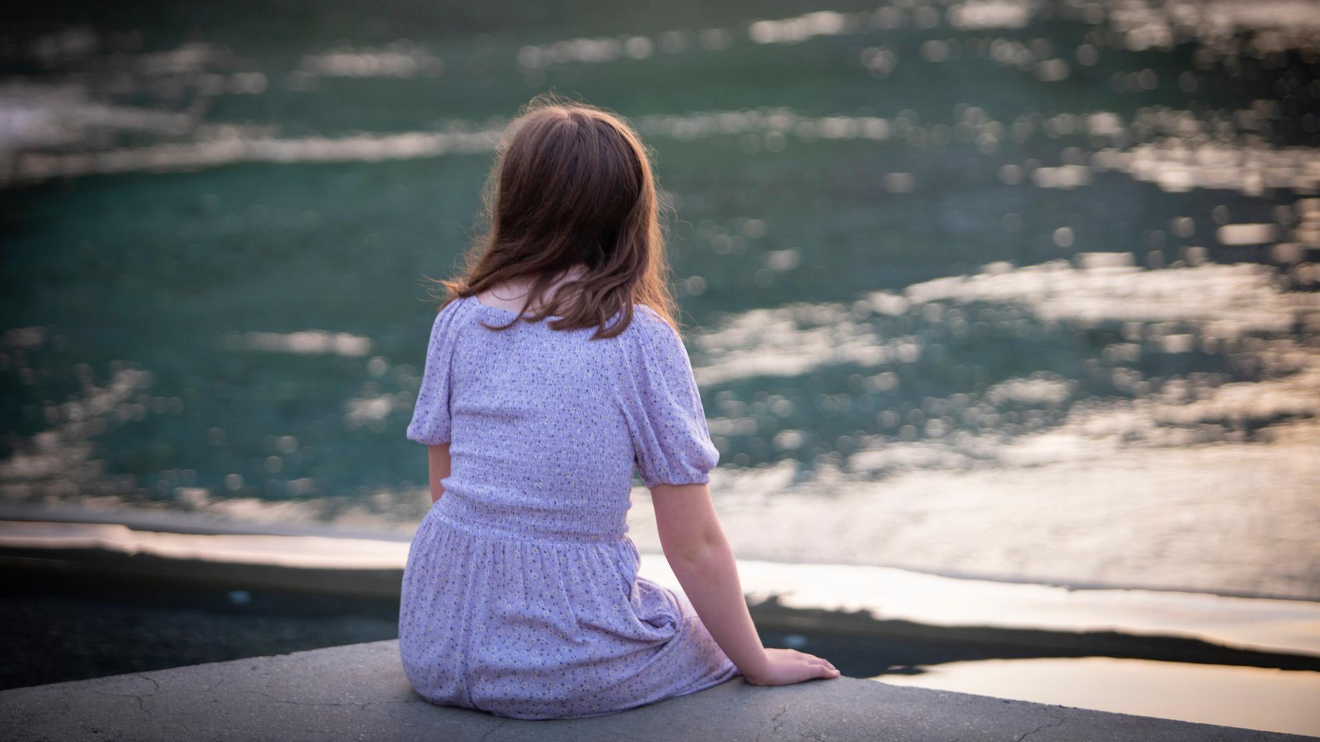 Ein Mädchen sitzt mit dem Rücken zur Kamera an einem Fluss-Ufer.